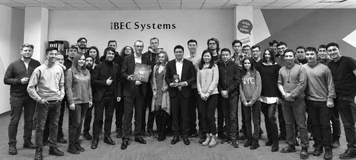 The iBEC Systems team standing together in the company office, under the iBEC Systems sign on the wall